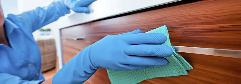 Cleaner wearing blue gloves wiping a wooden cabinet drawer with a microfiber cloth for dust and grime removal, Wilmington, NC.
