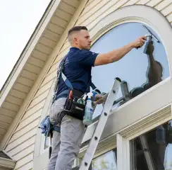 Professional window cleaner on a ladder using a squeegee to clean an exterior arched window, Wilmington, NC.